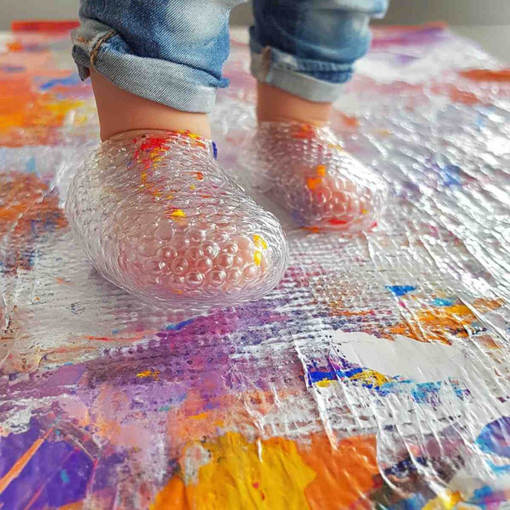 A toddler's feet wrapped in bubble wrap stepping on a colourful canvas, creating textured artwork. This image demonstrates an engaging 10-Minute Sensory Art Project using household items to encourage creativity and tactile exploration in young children.